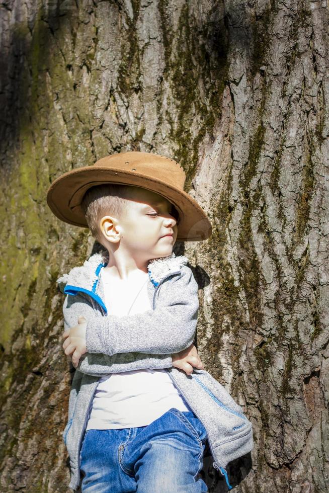 carino ragazzo in posa nel un' cowboy cappello nel il boschi di un' albero. il di sole raggi avvolgere il spazio. interazione storia per il libro. spazio per copiatura foto