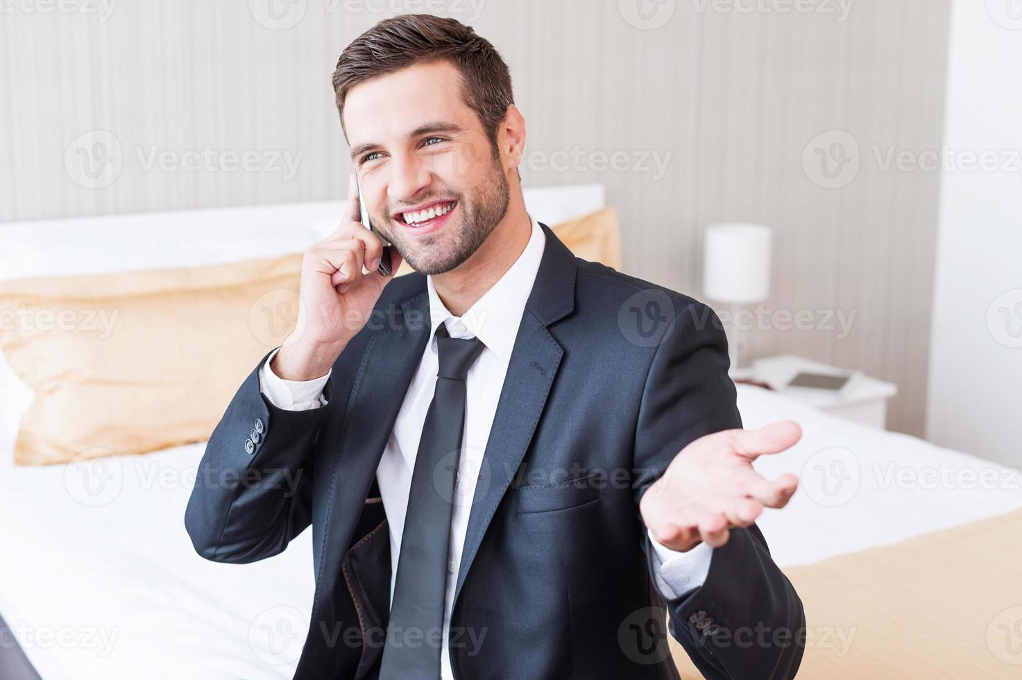 bene attività commerciale parlare. contento giovane uomo d'affari nel vestito formale parlando su il mobile Telefono e sorridente mentre seduta su il letto nel Hotel camera foto