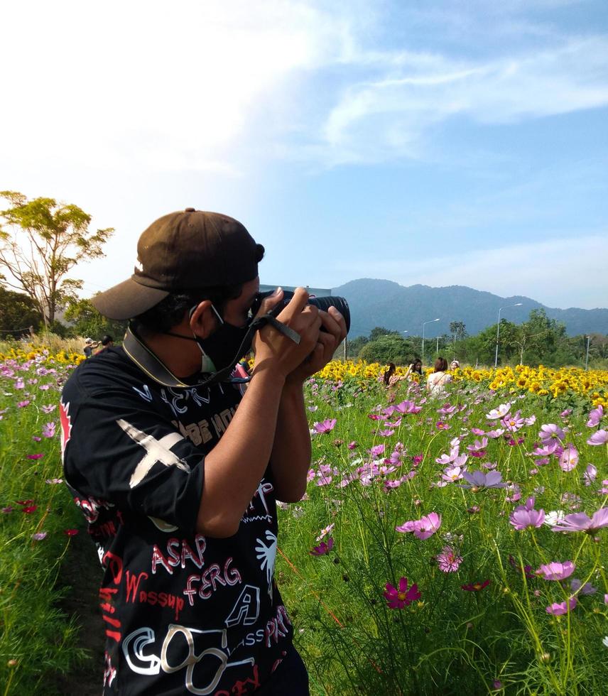 un' giovane fotografo indossare un' nero cappello e attrezzatura è assunzione immagini tra un' giardino di colorato fiori. luminosa cielo all'aperto foto