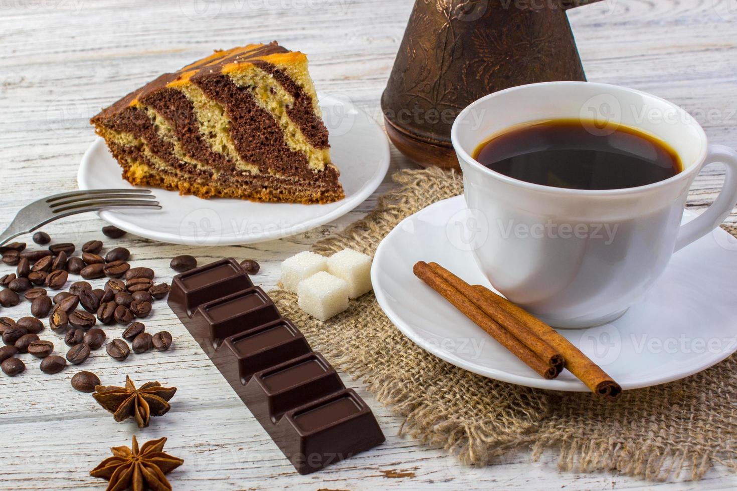 delizioso cioccolato torta su un' piatto con un' tazza di caffè . caffè fagioli, un' pezzo di cioccolato, un bel, zucchero e cannella bastoni su di legno tavolo foto