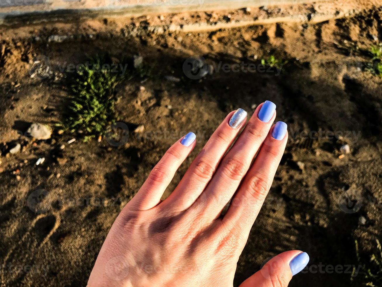 mano con blu manicure su sabbia sfondo. il squillare dito è dipinto nel argenteo paillettes. contro il sfondo di sporco e prato. costruzione di un' nuovo la zona, un' ragazza pittore Spettacoli corto Chiodi foto