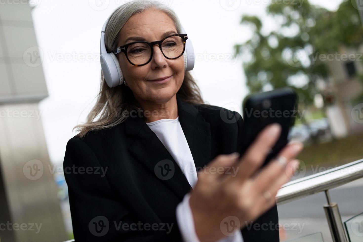 elegante anziano donna ascoltando per musica nel cuffie con un' Telefono nel sua mani foto