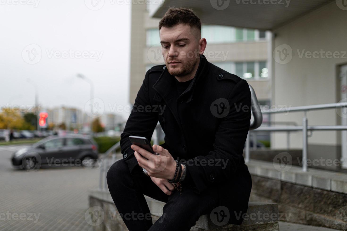 brutale giovane uomo nel un' nero cappotto con un' mobile Telefono su il sfondo di il ufficio centro foto
