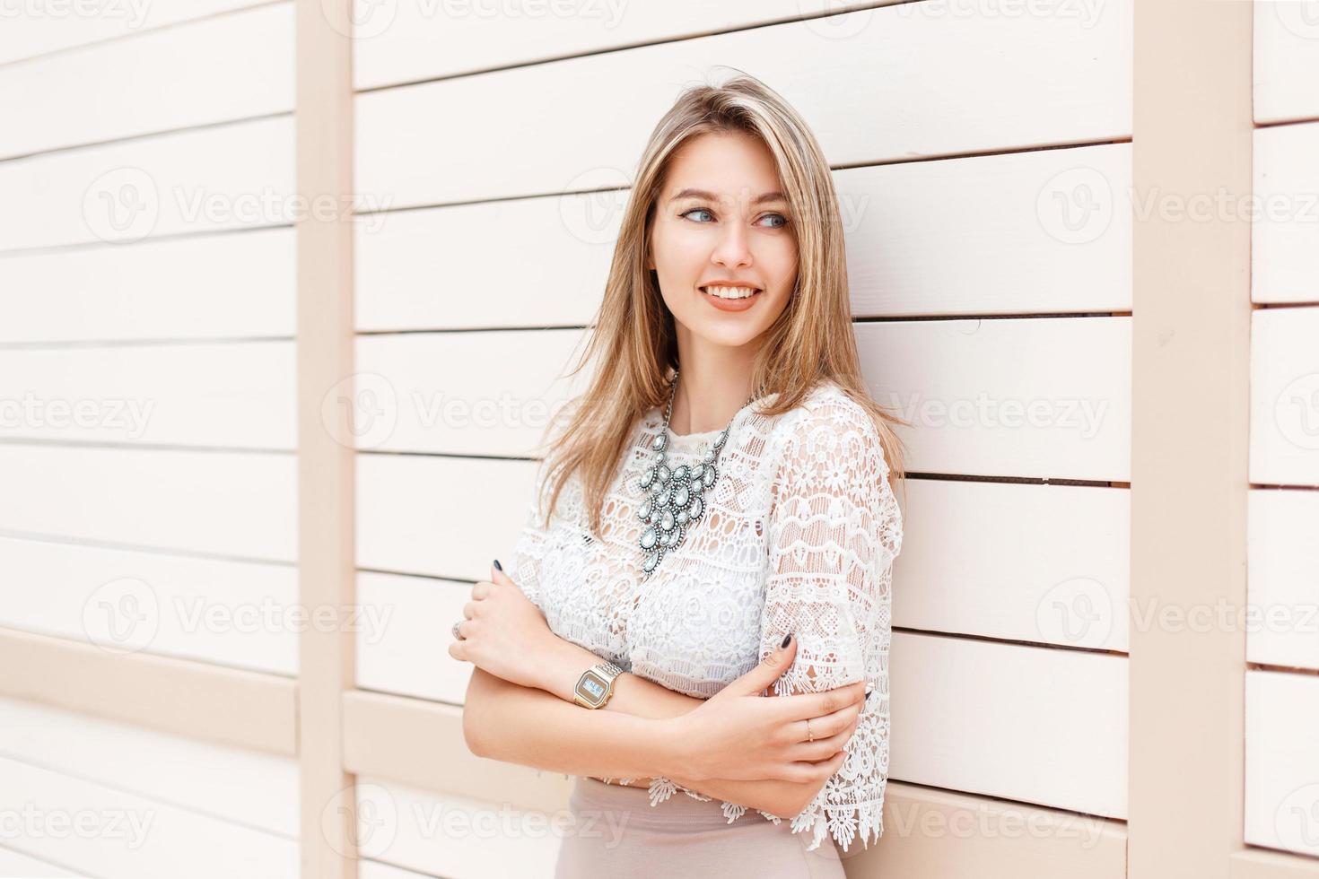 elegante bellissimo donna sorridente nel un' pizzo camicetta e vestito sta vicino un' bianca di legno parete. foto