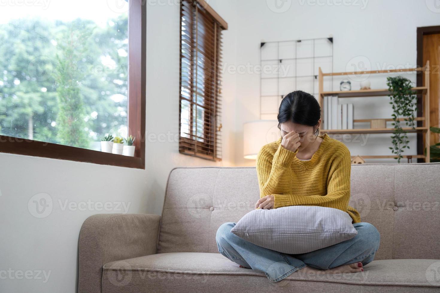 mano su templi di giovane infelice tristezza asiatico ragazza seduta su divano. lei è sensazione non molto bene dovuto per sua malattia e avendo un' male alla testa. visto a partire dal il davanti. fatica e malattia concetto. foto