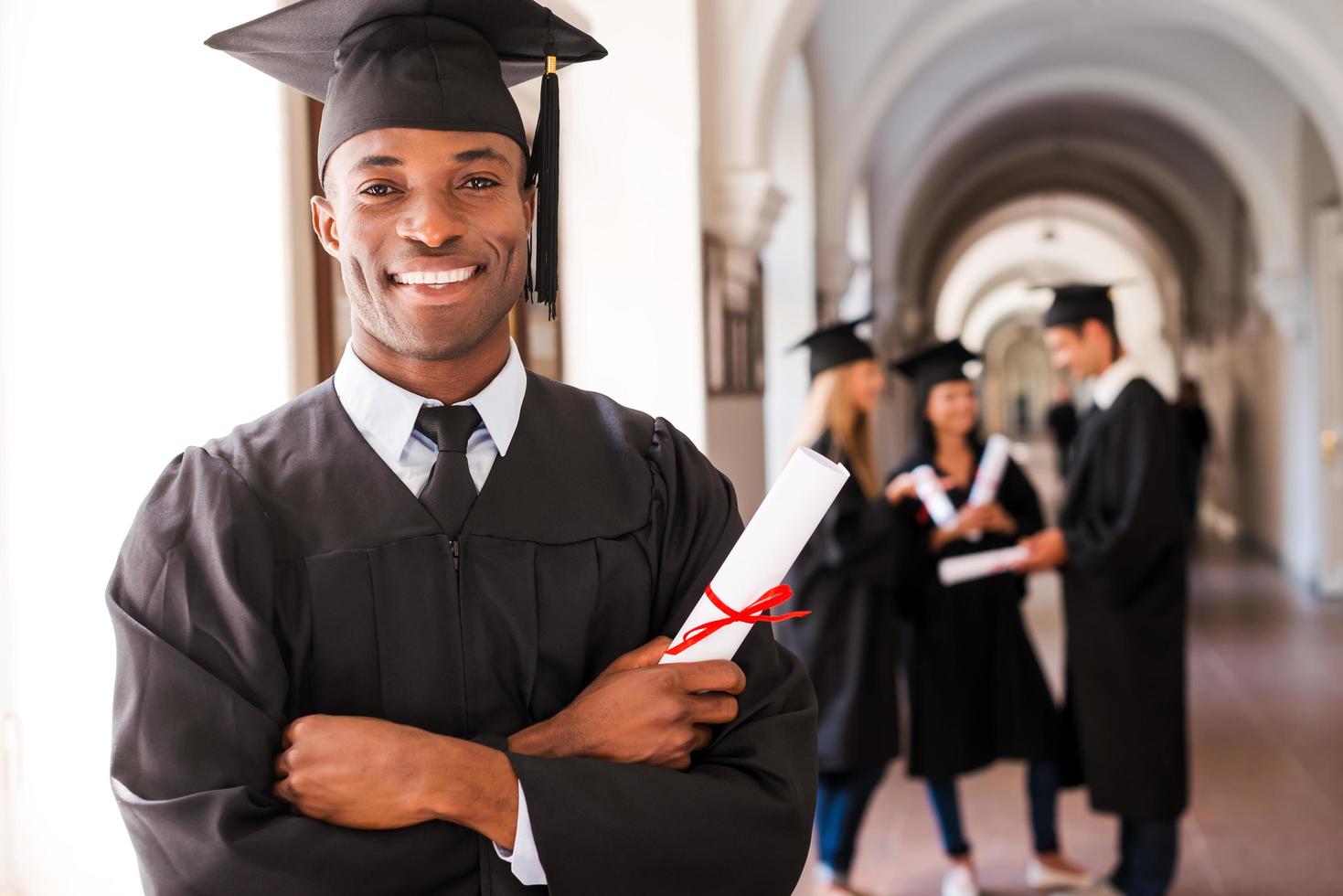 fiducioso laureato. contento africano uomo nel la laurea abiti Tenere diploma e sorridente mentre il suo amici in piedi nel il sfondo foto