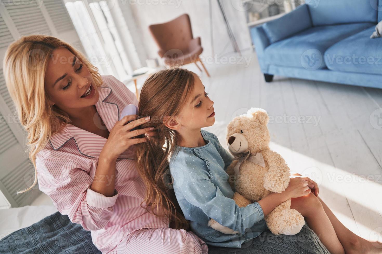 Perfetto parrucchiere. superiore Visualizza di bellissimo giovane madre spazzolatura sua figlia capelli mentre seduta su il letto a casa foto