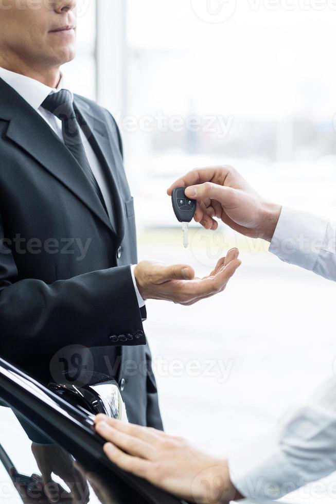 dando un' chiave per un' nuovo auto proprietario. ritagliata Immagine di fiducioso uomo nel vestito formale ricevente un' chiave a partire dal il suo nuovo auto foto