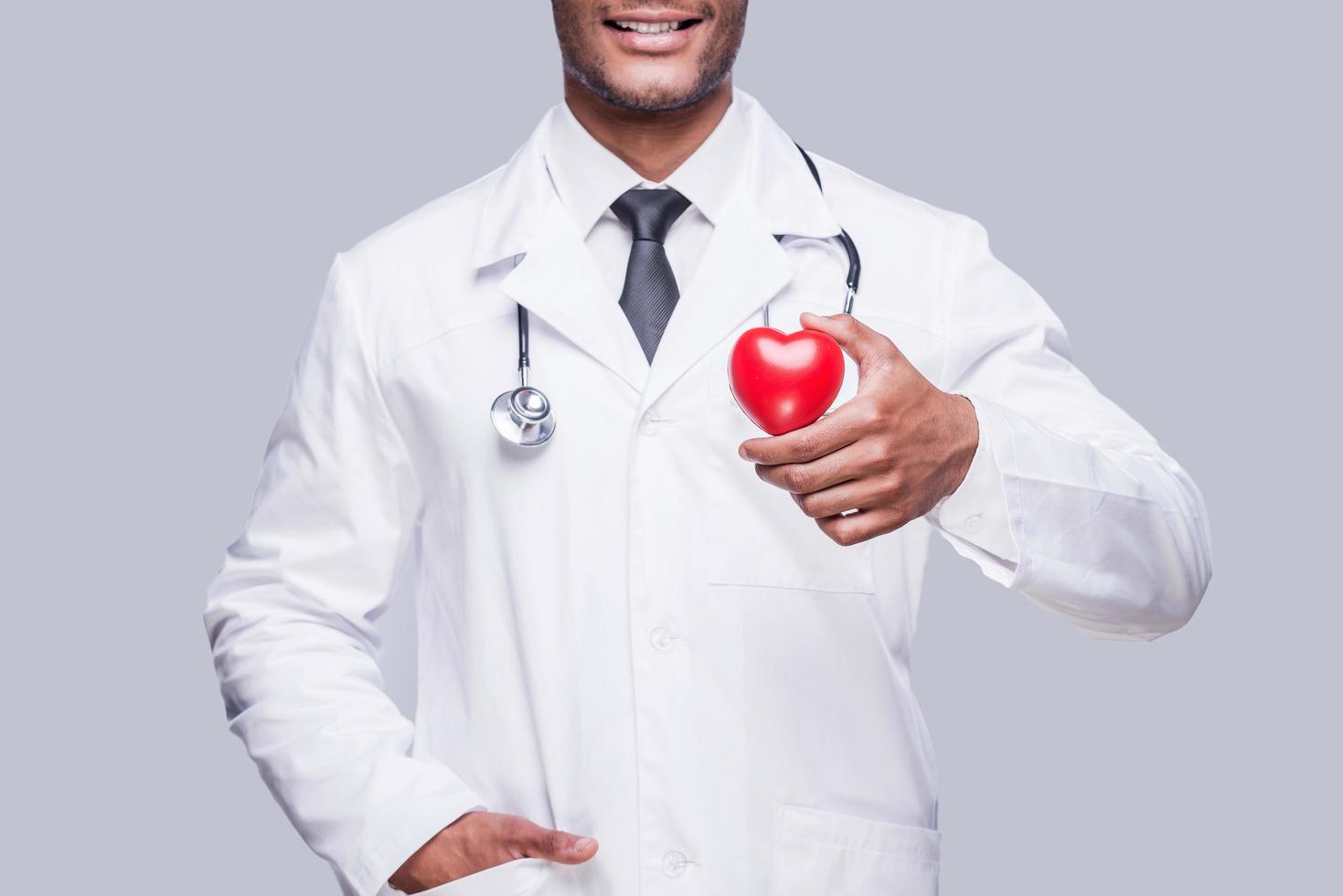 assunzione cura di il tuo cuore. ritagliata Immagine di fiducioso africano medico Tenere cuore forma giocattolo e sorridente mentre in piedi contro grigio sfondo foto