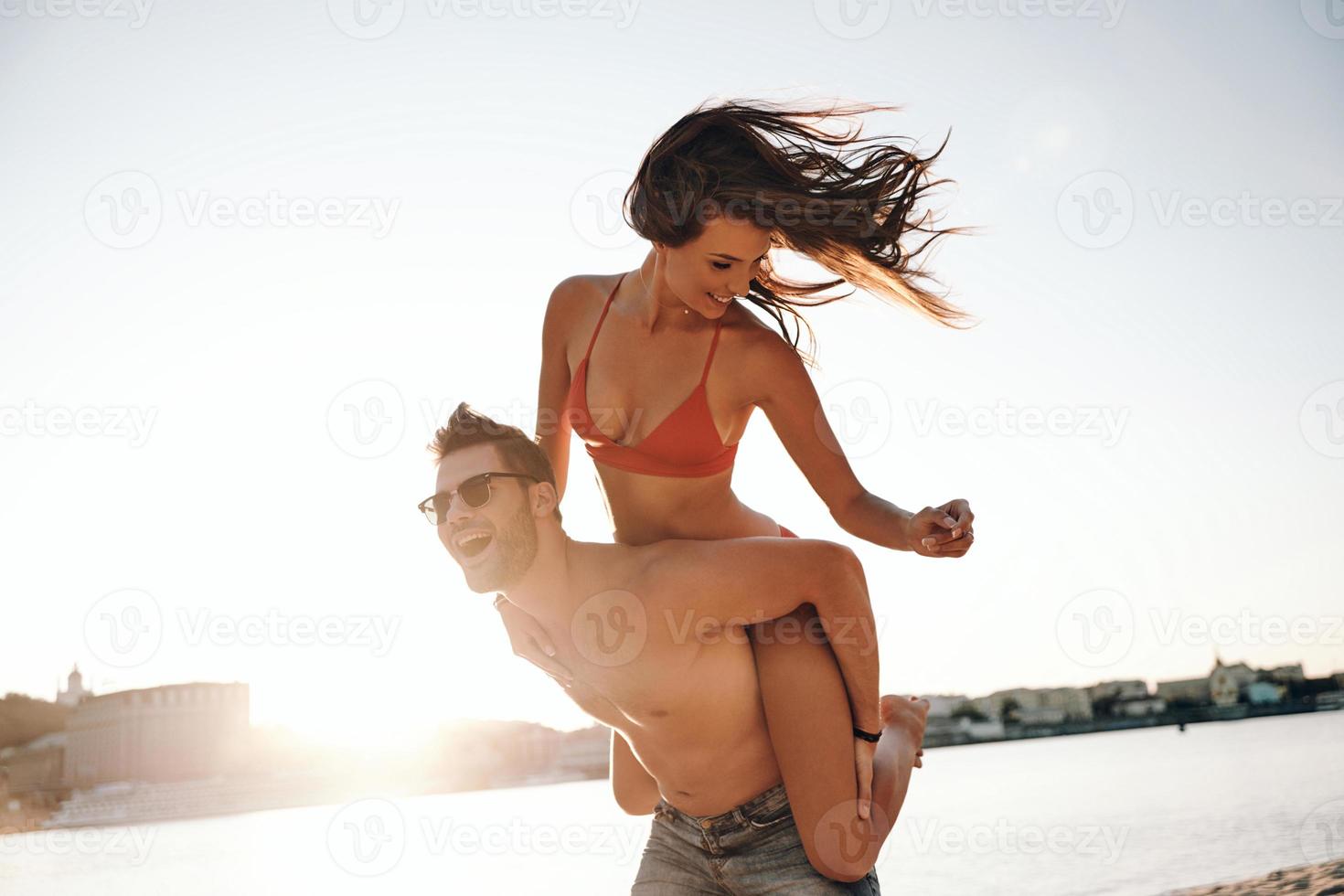 appena avendo divertimento. bello giovane uomo dando il suo attraente fidanzata un' porcellino indietro cavalcata mentre a piedi su il spiaggia foto