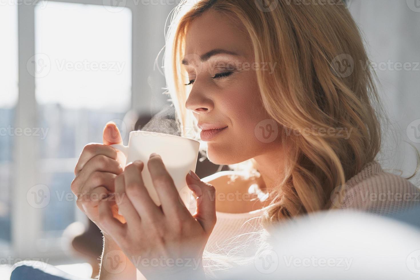 semplicemente contento. attraente giovane donna Tenere un' tazza e conservazione occhi chiuso con Sorridi mentre la spesa tempo a casa foto