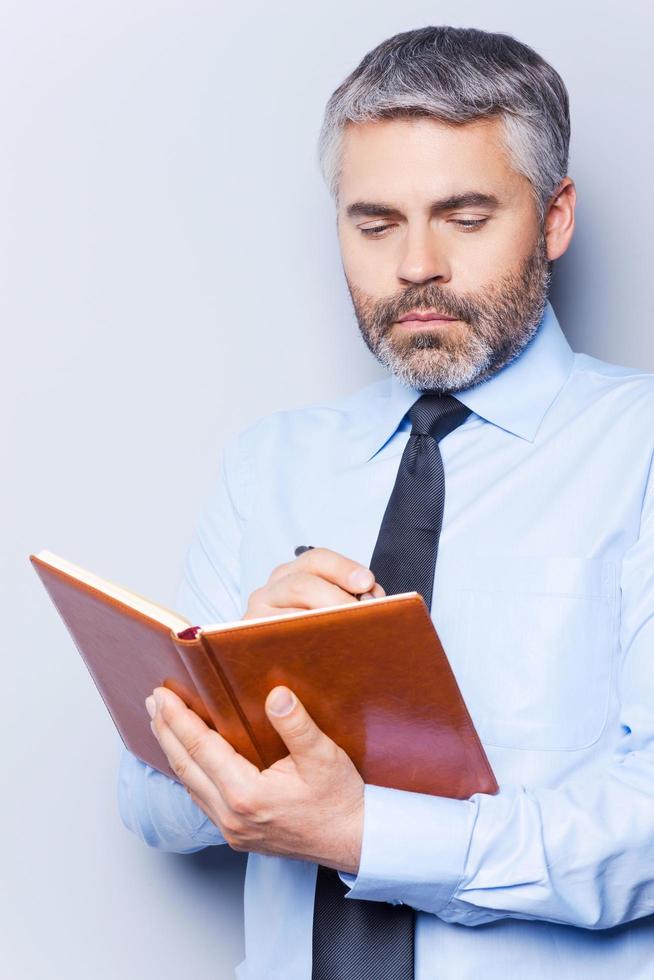 scrittura fare elenco. fiducioso maturo uomo nel camicia e cravatta scrittura qualcosa nel Nota tampone mentre in piedi contro grigio sfondo foto