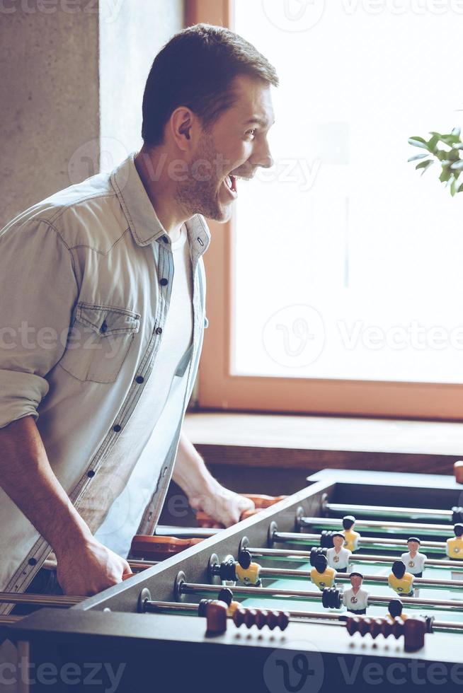 amorevole Questo gioco. lato Visualizza di allegro giovane bello uomo giocando Foosball gioco e guardare eccitato mentre in piedi nel davanti di finestra foto