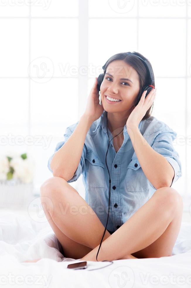 ascoltando per sua preferito musica. allegro giovane donna nel cuffie ascoltando per mp3 giocatore e sorridente a telecamera mentre seduta nel letto foto