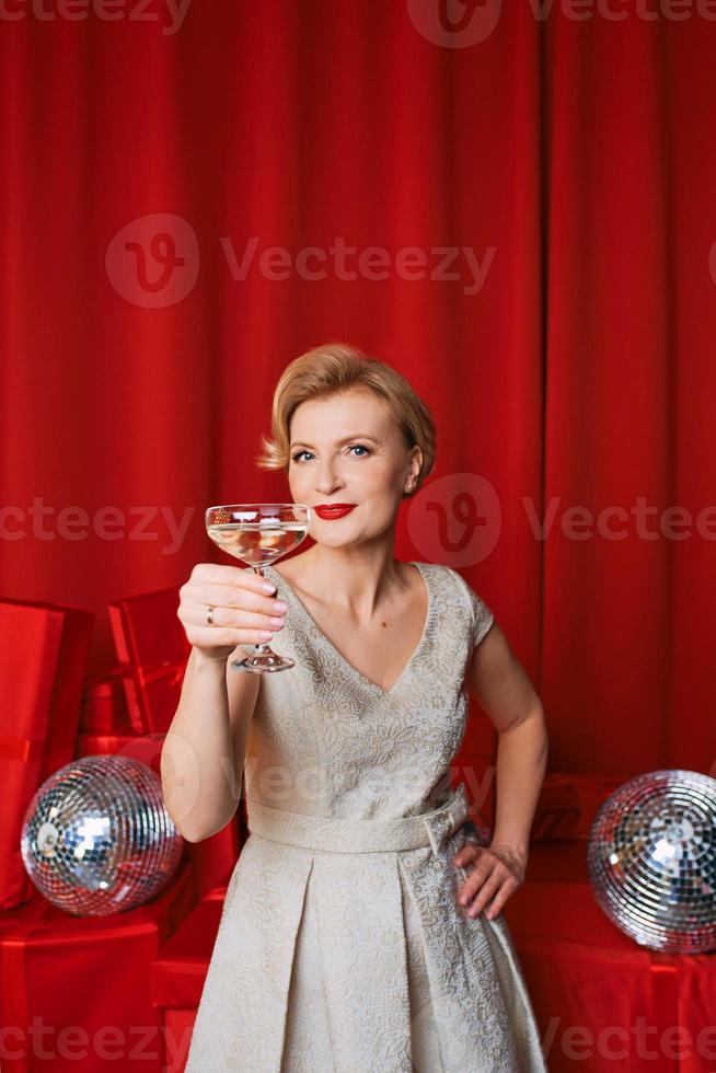 maturo elegante elegante donna nel cocktail vestito con bicchiere di scintillante vino con regali su rosso sfondo. festa, moda, celebrazione, anti età concetto foto