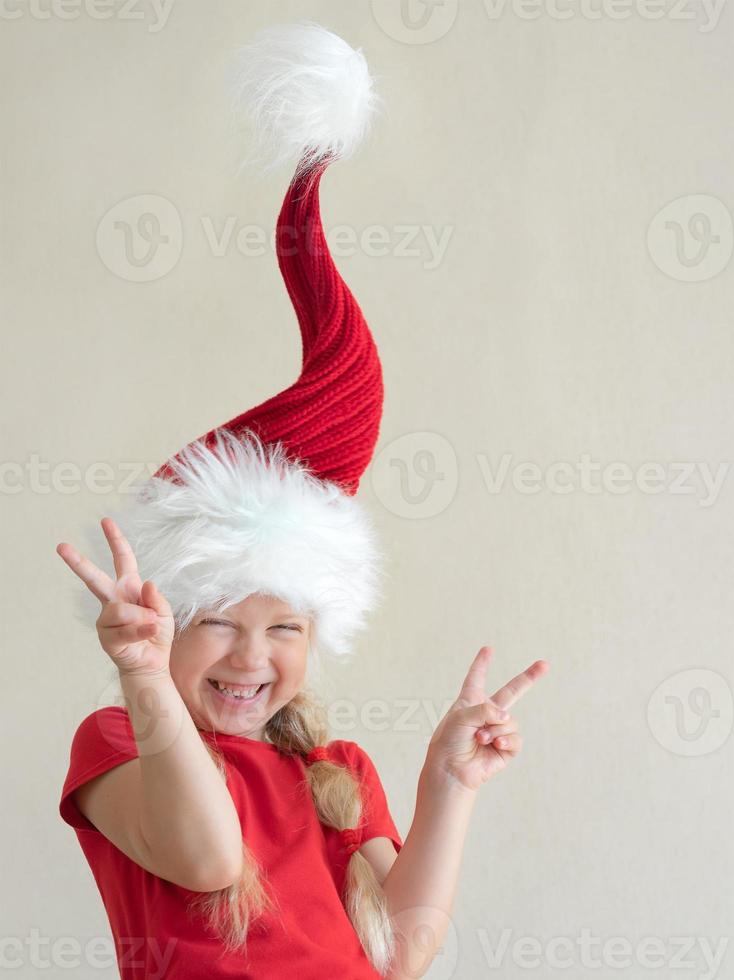 divertente poco ragazza nel il Santa cappello, quello in piedi su, sorridente e mostrare Due vittoria con sua dita foto