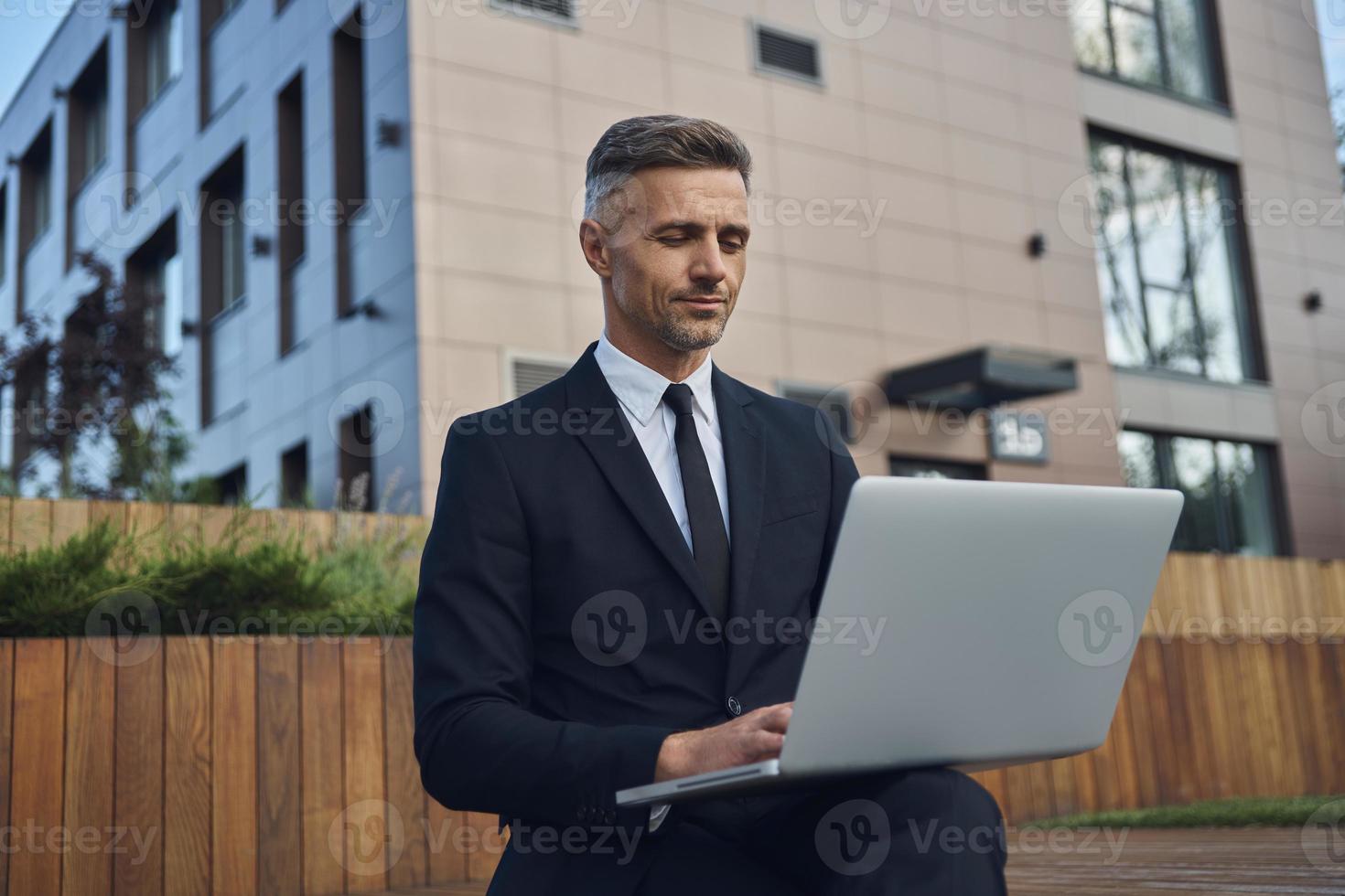 fiducioso maturo uomo d'affari Lavorando su il computer portatile mentre seduta all'aperto vicino ufficio edificio foto