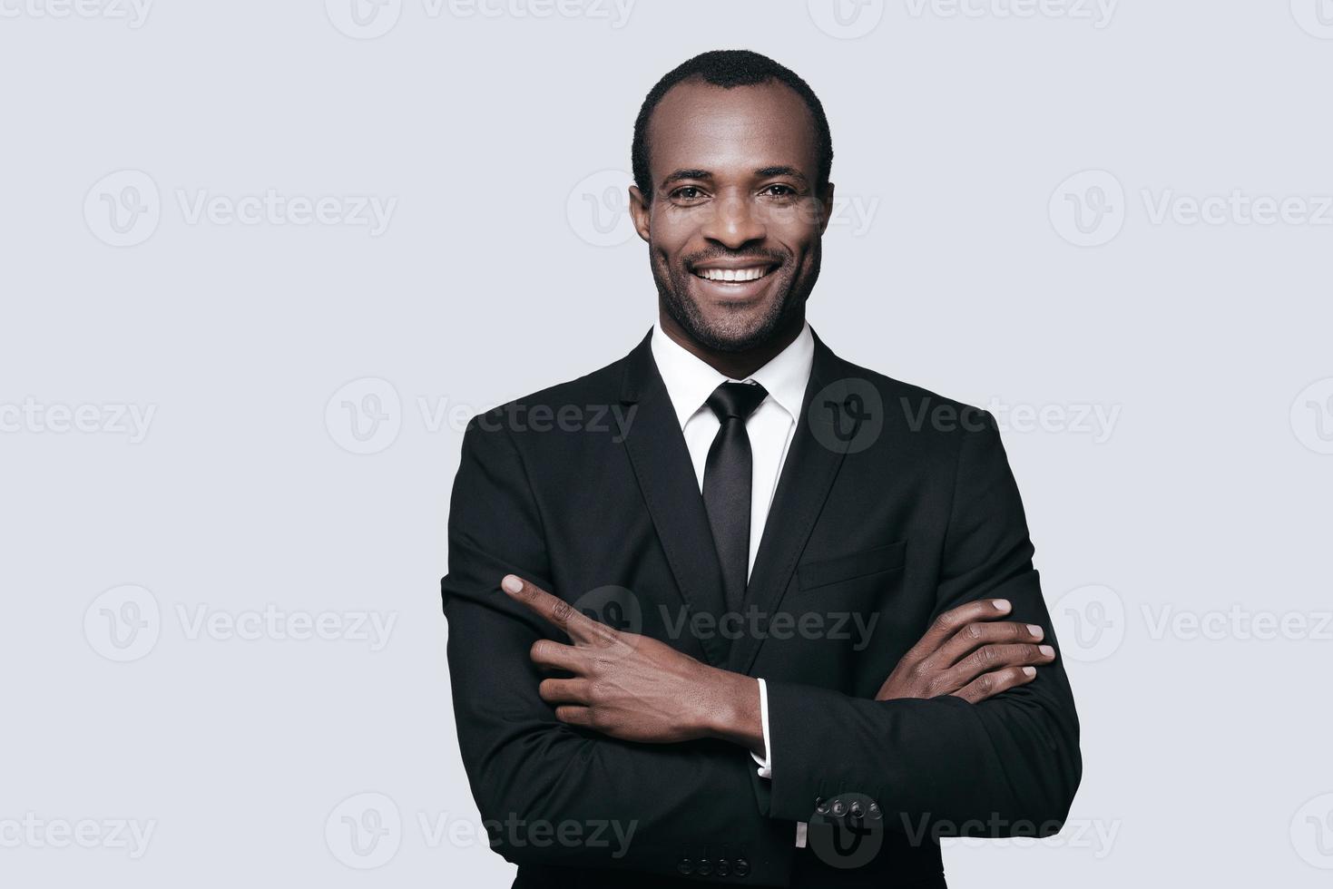 puntamento in qualche luogo. bello giovane africano uomo nel vestito formale puntamento da qualche parte e sorridente mentre in piedi contro grigio sfondo foto