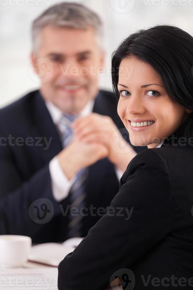 donna su colloquio. allegro giovane donna nel vestito formale guardare al di sopra di spalla e sorridente mentre maturo uomo nel completo da uomo seduta su sfondo foto