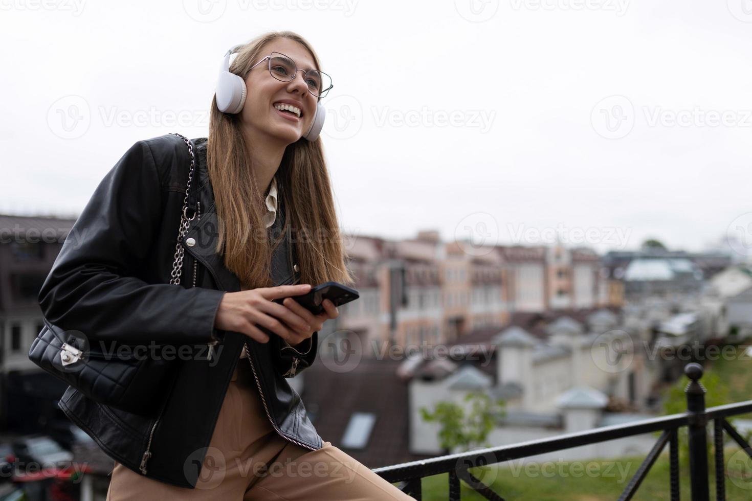 un' giovane alunno ascolta per piacevole musica contro il fondale di il urbano paesaggio nel cuffie con un' Telefono nel sua mani foto