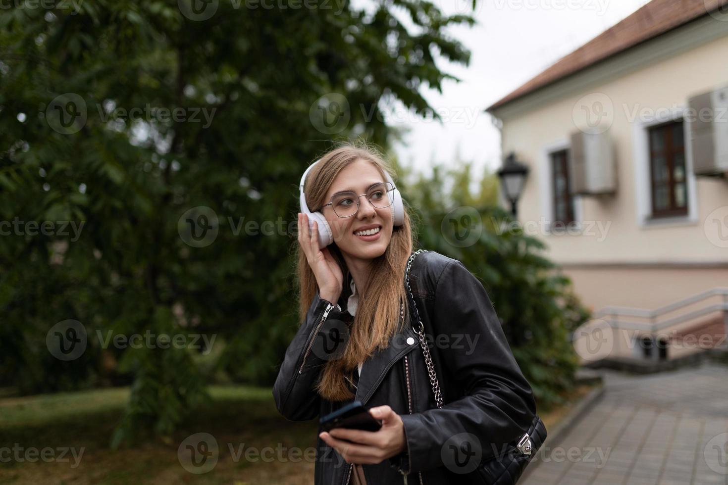 sorridente giovane femmina alunno ascoltando per musica con cuffie su il sfondo di il urbano paesaggio foto