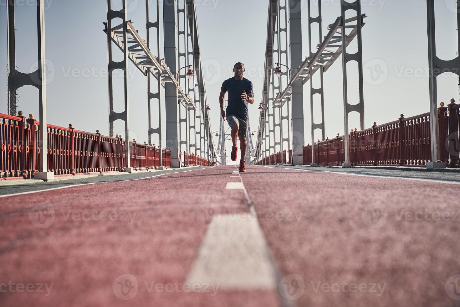 sempre nel grande forma. pieno lunghezza di giovane africano uomo nel gli sport capi di abbigliamento esercizio mentre jogging su il ponte all'aperto foto