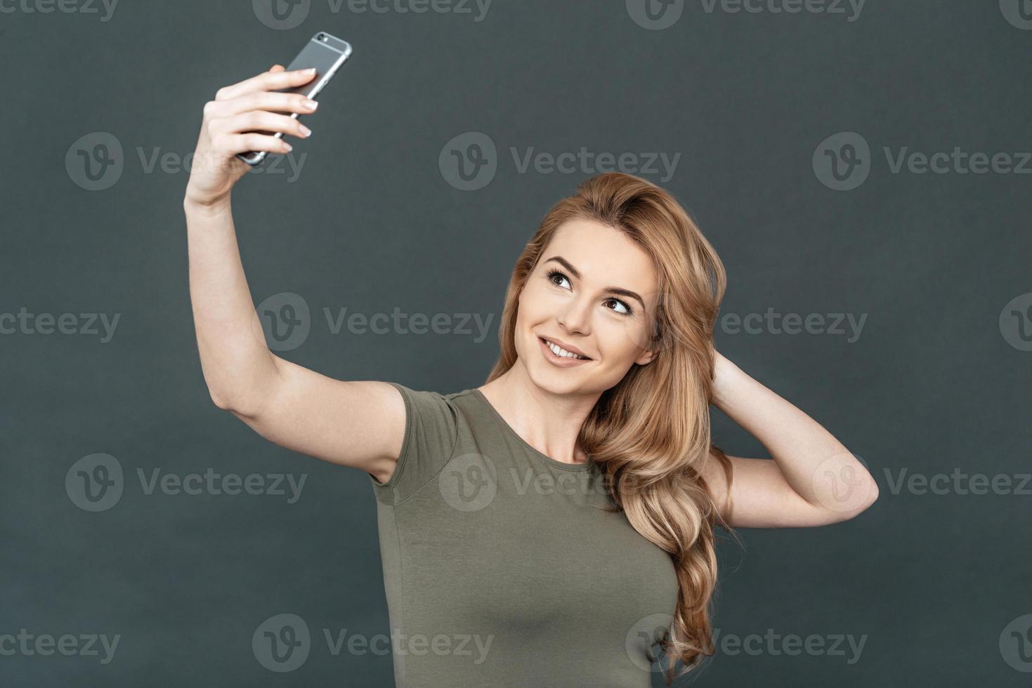 assunzione autoscatto. ritratto di attraente giovane sorridente donna con biondo capelli fabbricazione autoscatto di sua inteligente Telefono mentre in piedi contro grigio sfondo foto