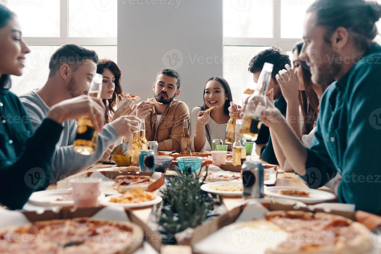 grande festa. gruppo di giovane persone nel casuale indossare mangiare Pizza e sorridente mentre avendo un' cena festa in casa foto