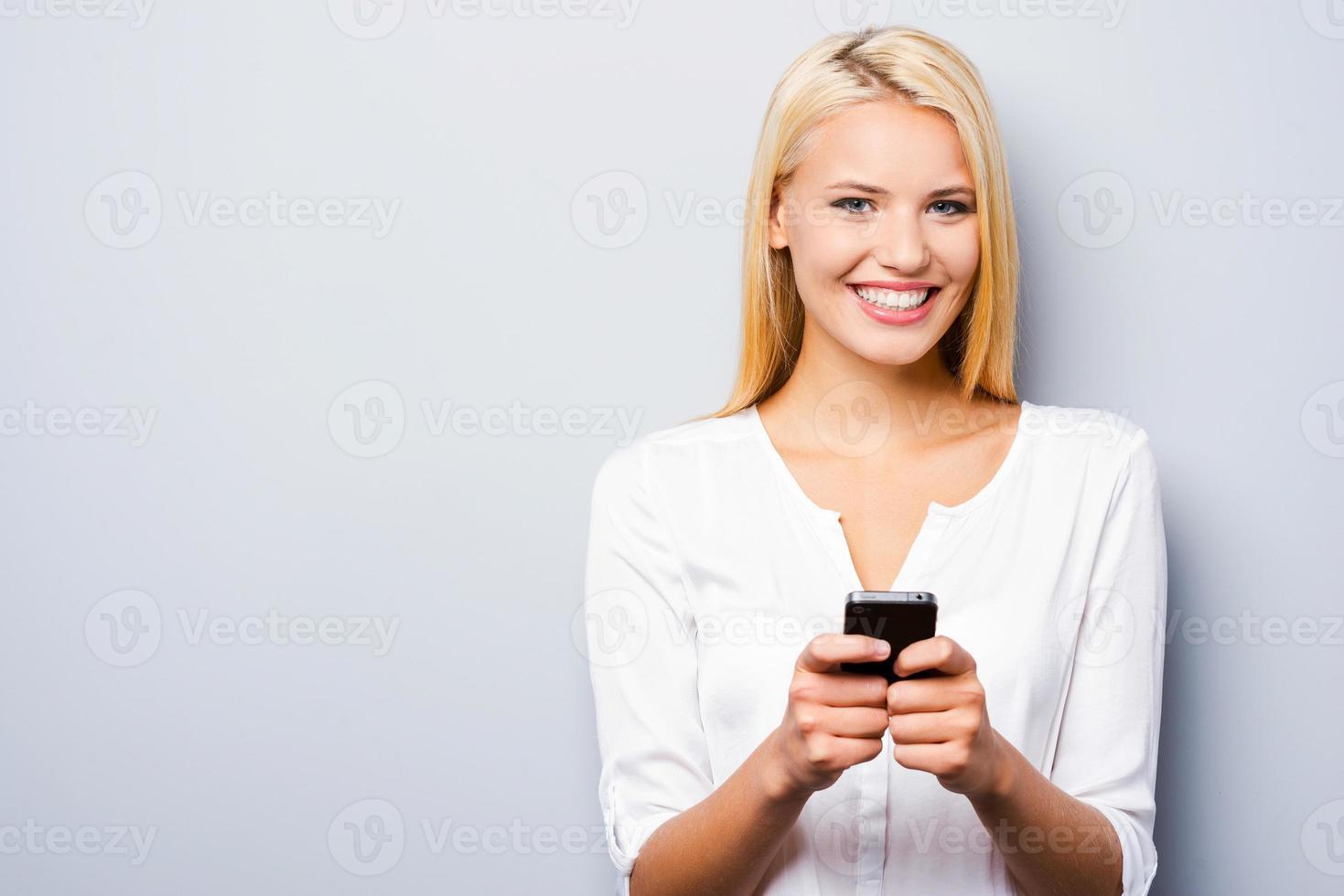 l'esame sua marca nuovo inteligente Telefono. bellissimo giovane biondo capelli donne Tenere inteligente Telefono e sorridente mentre in piedi contro grigio sfondo foto