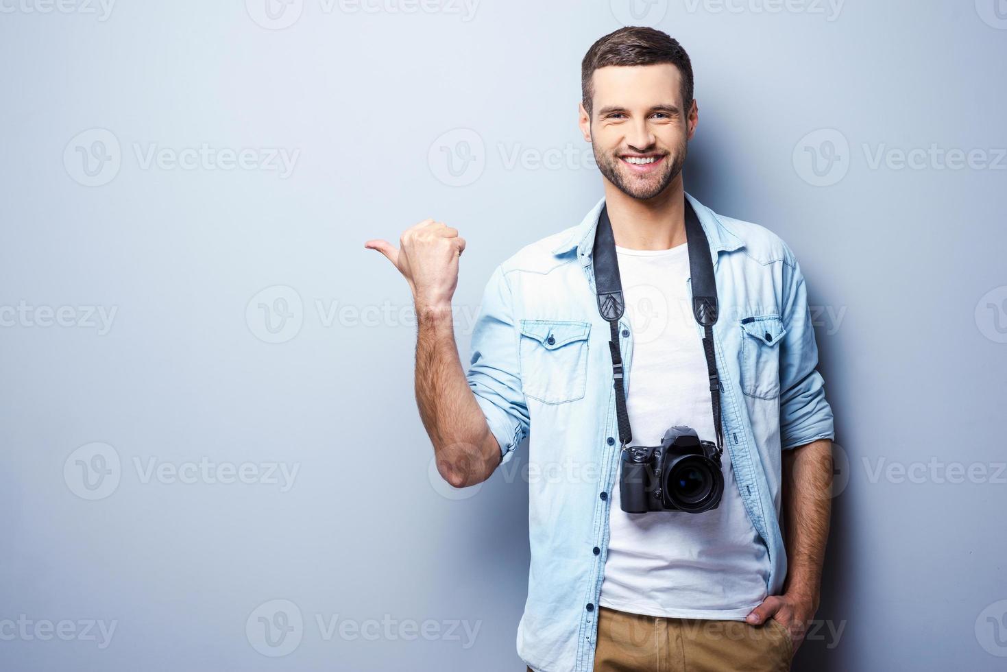 fotografi la scelta esso. bello giovane uomo con digitale telecamera puntamento lontano e sorridente mentre in piedi contro grigio sfondo foto