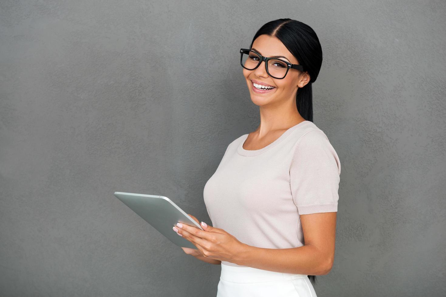 lei conosce il segreto di successo. allegro giovane donna d'affari Tenere digitale tavoletta e guardare a telecamera mentre in piedi contro grigio sfondo foto