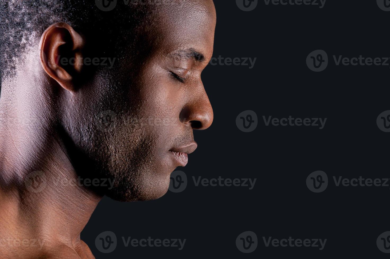 calma e fiducioso. lato Visualizza di giovane africano uomo conservazione occhi chiuso mentre in piedi contro grigio sfondo foto