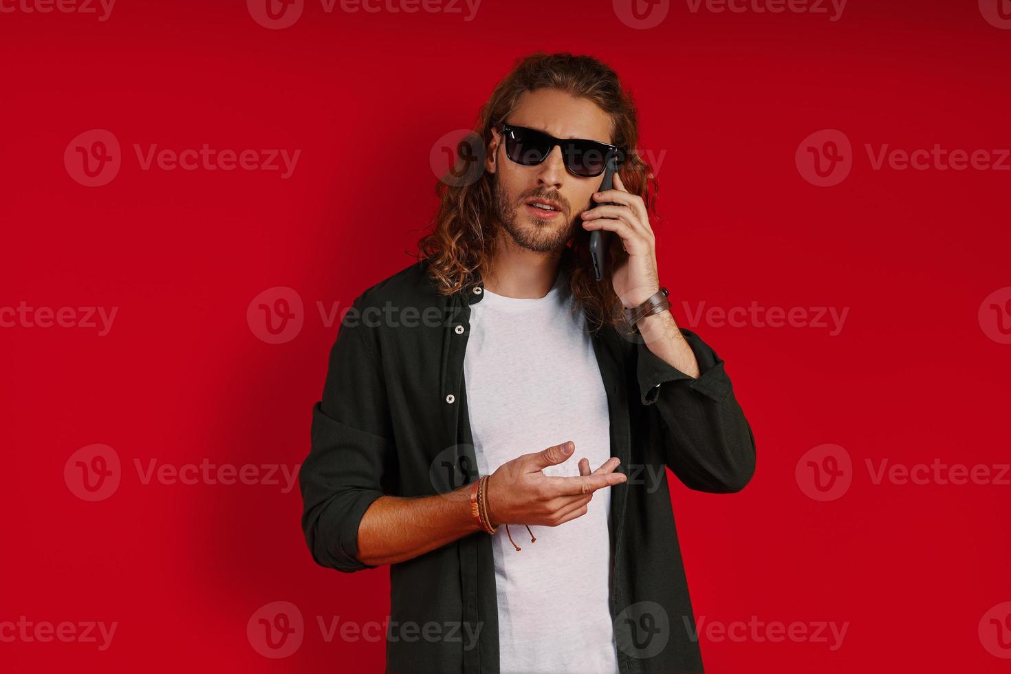 occupato giovane uomo nel casuale capi di abbigliamento guardare a telecamera e parlando su il Telefono mentre in piedi contro rosso sfondo foto
