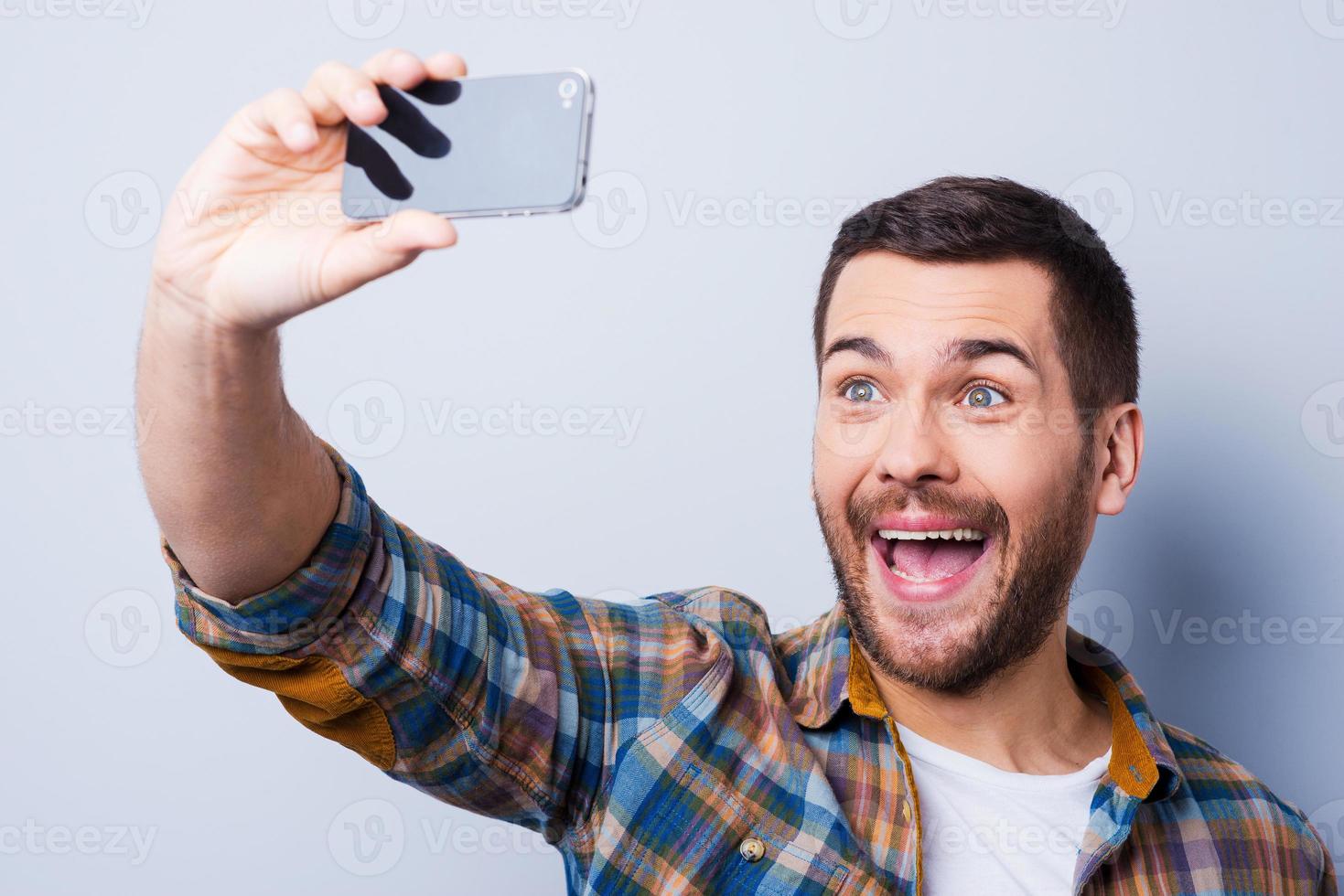 allegro autoscatto. allegro giovane uomo nel camicia Tenere mobile Telefono e fabbricazione foto di lui stesso mentre in piedi contro grigio sfondo