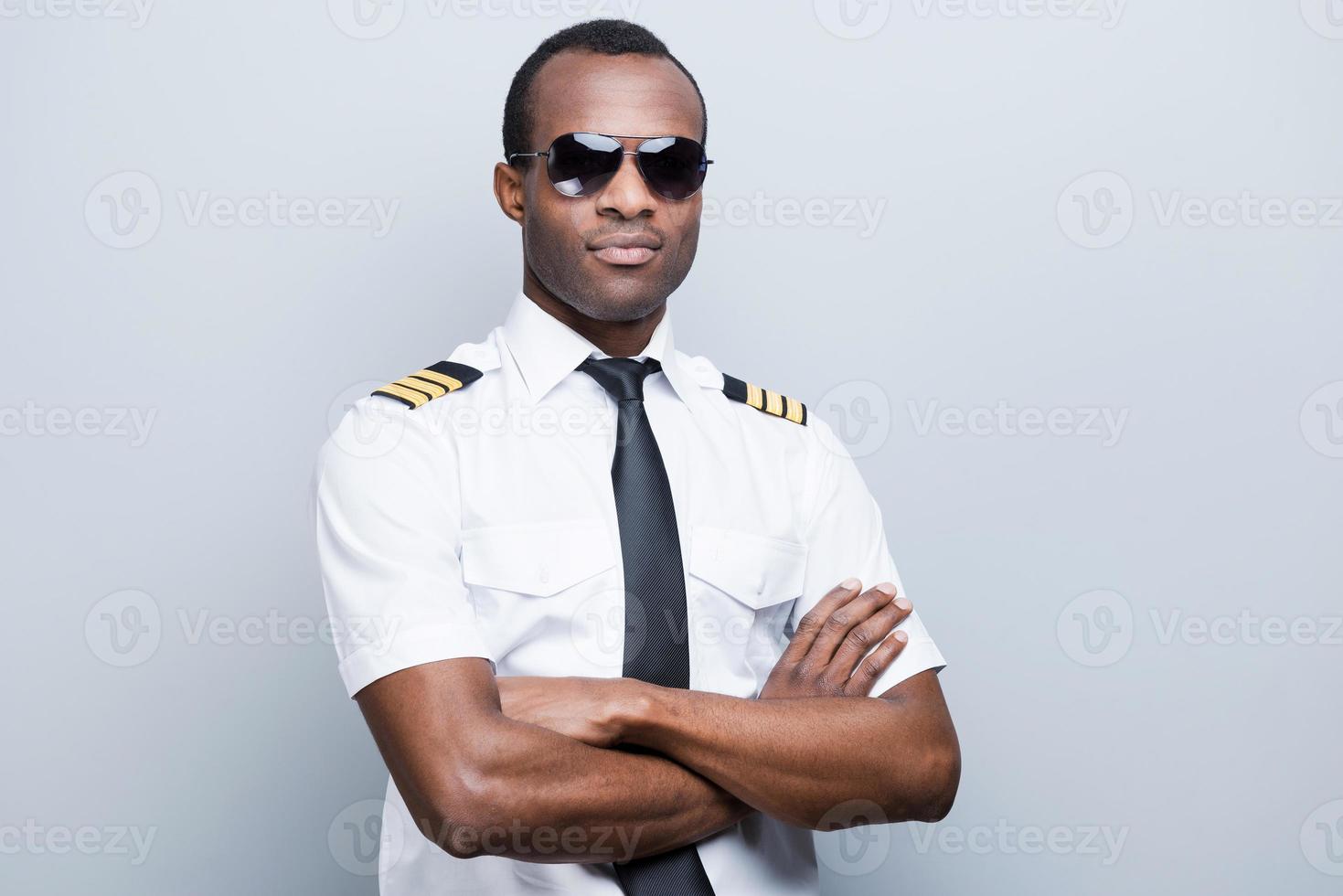 fiducioso e esperto pilota. fiducioso africano pilota nel uniforme conservazione braccia attraversato mentre in piedi contro grigio sfondo foto