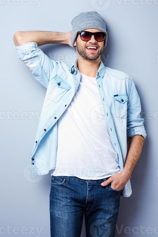 giovane e freddo. bello giovane elegante uomo nel occhiali da sole e cappello guardare a telecamera e sorridente mentre in piedi contro grigio sfondo foto