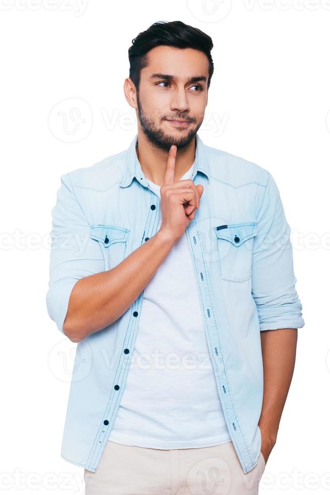 nel ricerca di soluzione. riflessivo giovane indiano uomo toccante il suo mento con dito e guardare lontano mentre in piedi contro bianca sfondo foto