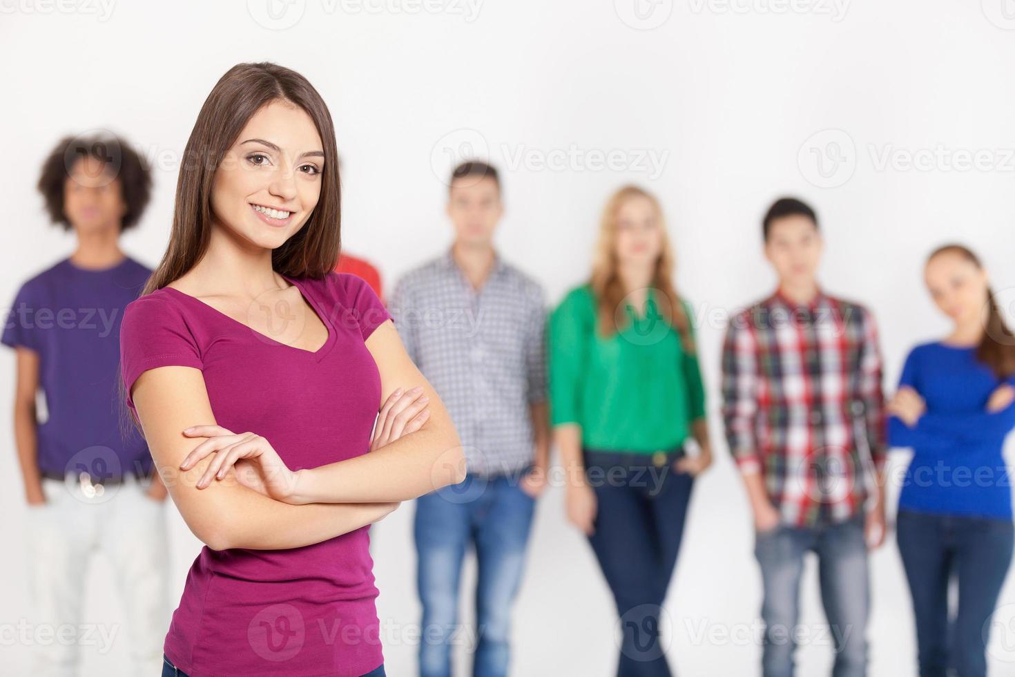 fiducioso nel sua bellezza. allegro giovane donna conservazione braccia attraversato e sorridente mentre sua amici in piedi su sfondo foto