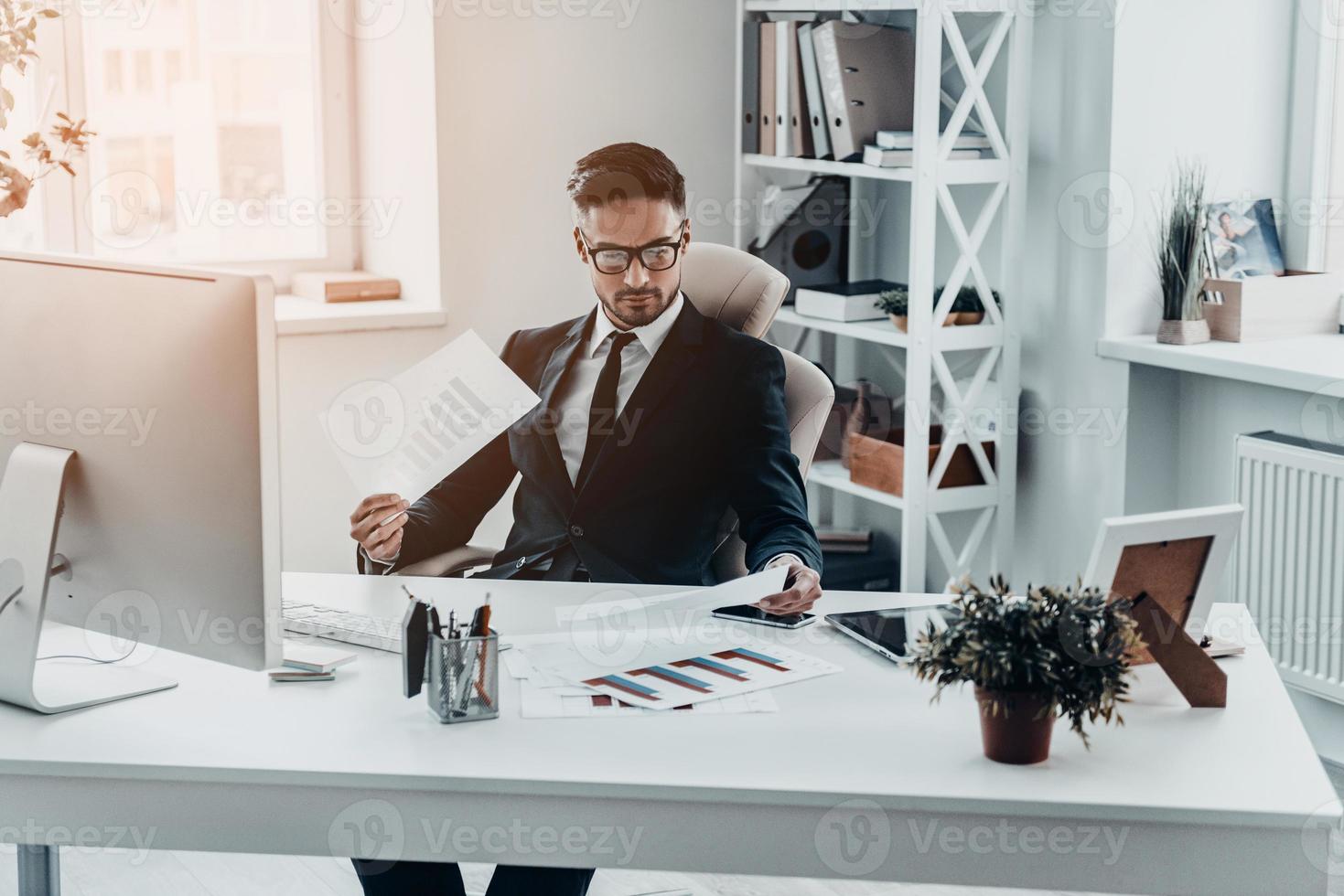 controllo il rapporti. bello giovane uomo nel pieno completo da uomo Lavorando con documenti mentre seduta a il ufficio scrivania foto
