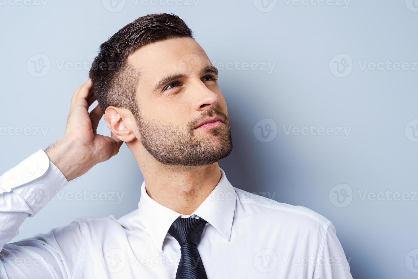 pensiero di soluzioni. riflessivo giovane uomo nel camicia e cravatta toccante testa con mano e guardare lontano mentre in piedi contro grigio sfondo foto