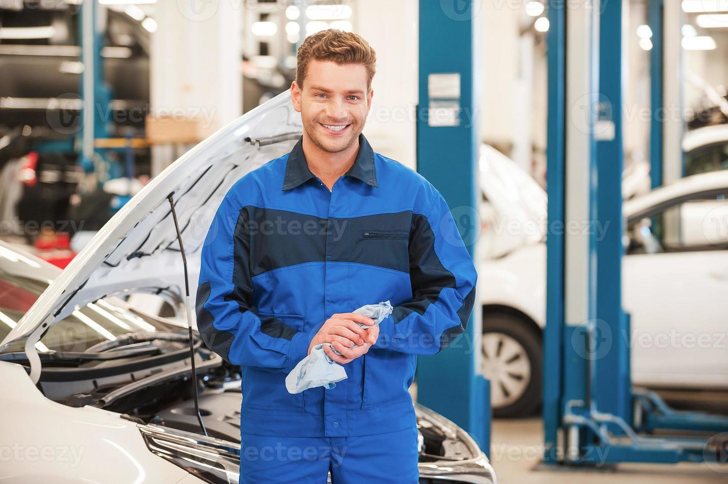 dopo il opera è fatto. fiducioso giovane uomo nel uniforme asciugandosi il suo mani con straccio e sorridente mentre in piedi nel laboratorio con auto nel il sfondo foto