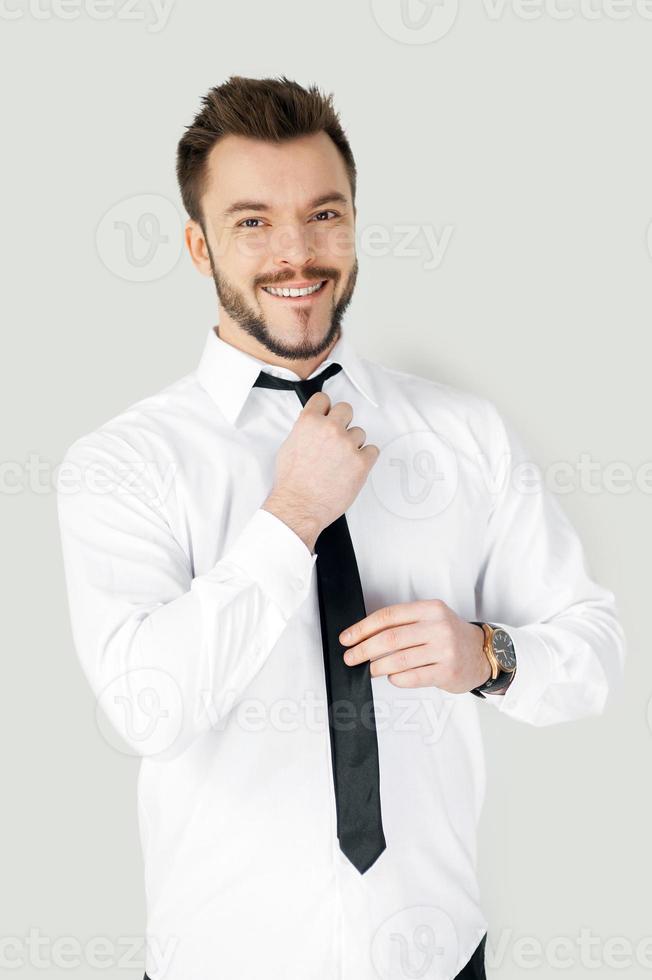 attività commerciale condimento su. bello giovane uomo nel vestito formale regolazione il suo cravatta e guardare a telecamera mentre in piedi contro grigio sfondo foto