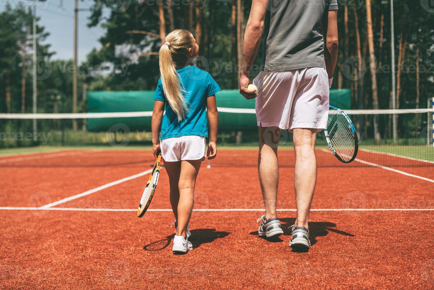 preparazione per grande gioco. posteriore Visualizza di poco biondo capelli ragazza nel gli sport capi di abbigliamento trasporto tennis racchetta e guardare a sua padre a piedi vicino sua di tennis Tribunale foto