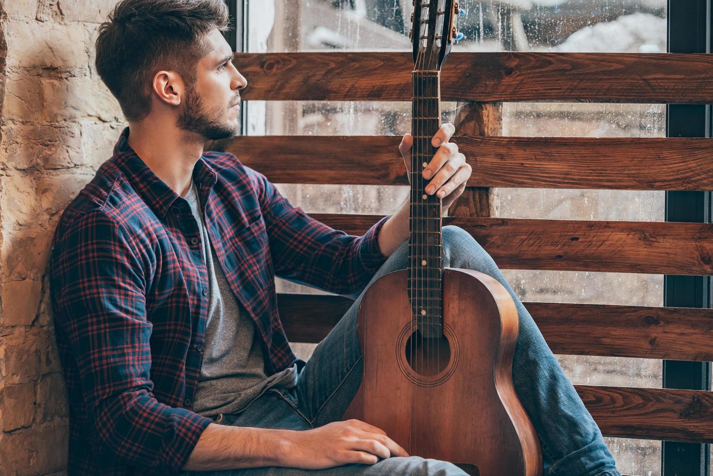 musicale ispirazioni. lato Visualizza di bello giovane uomo Tenere chitarra e guardare attraverso finestra mentre seduta a davanzale foto