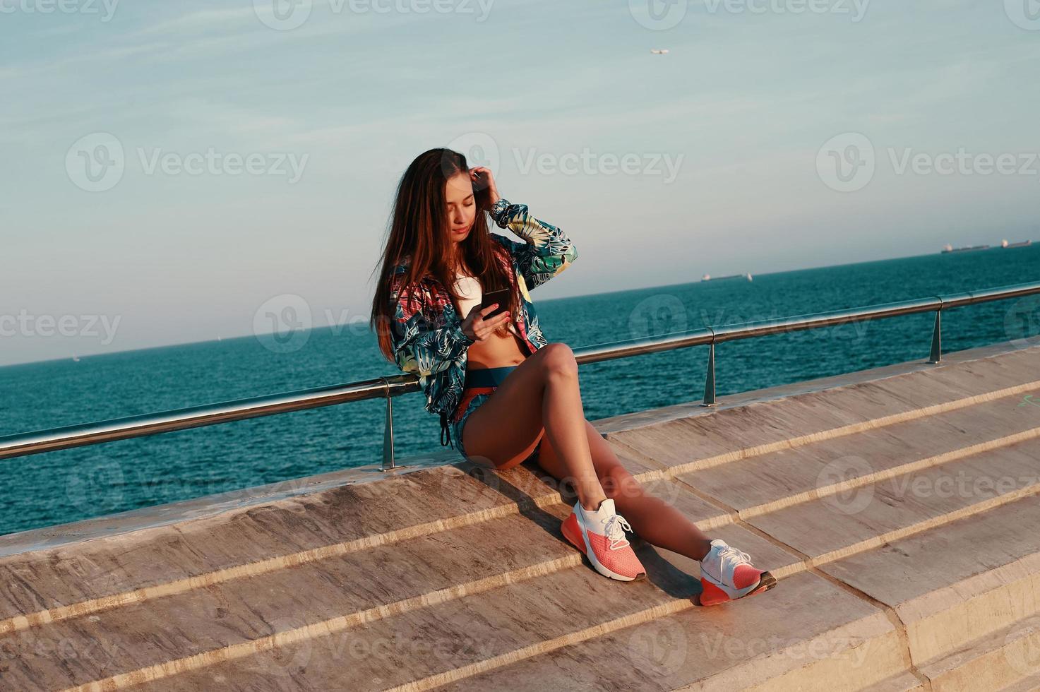 utilizzando moderno tecnologie. attraente giovane donna utilizzando inteligente Telefono mentre seduta vicino il spiaggia all'aperto foto