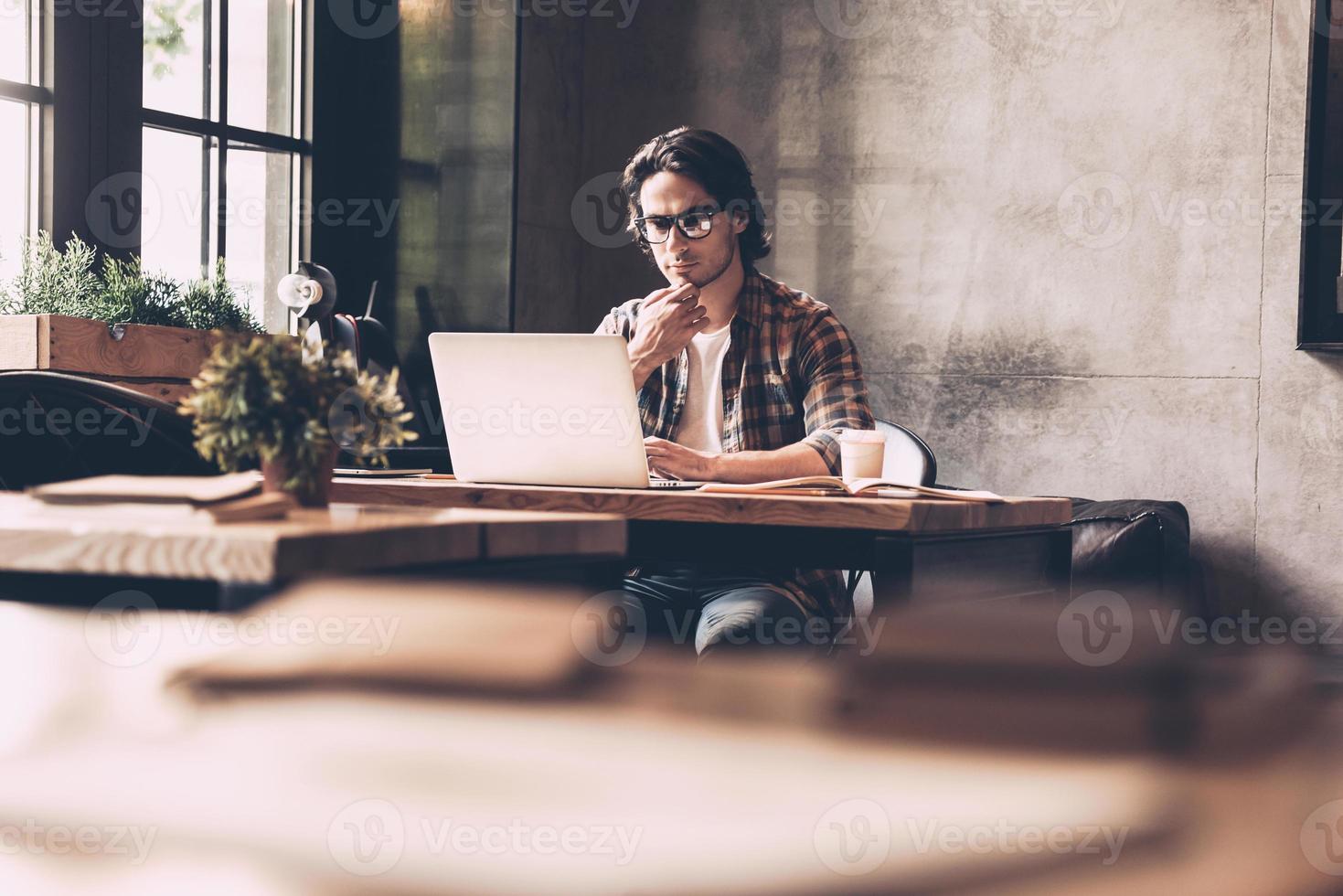 uomo d'affari a opera. fiducioso giovane uomo nel casuale indossare guardare a il computer portatile e Tenere mano su mento mentre seduta vicino finestra nel creativo ufficio foto