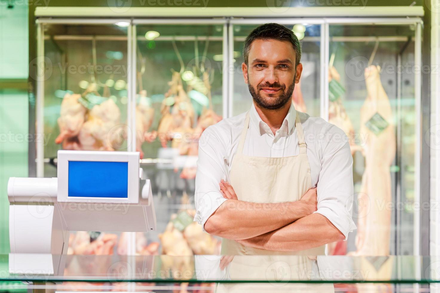 fiducioso macellaio. sorridente giovane macellaio conservazione braccia attraversato e guardare a telecamera mentre in piedi a supermercato check-out con carne nel il sfondo foto