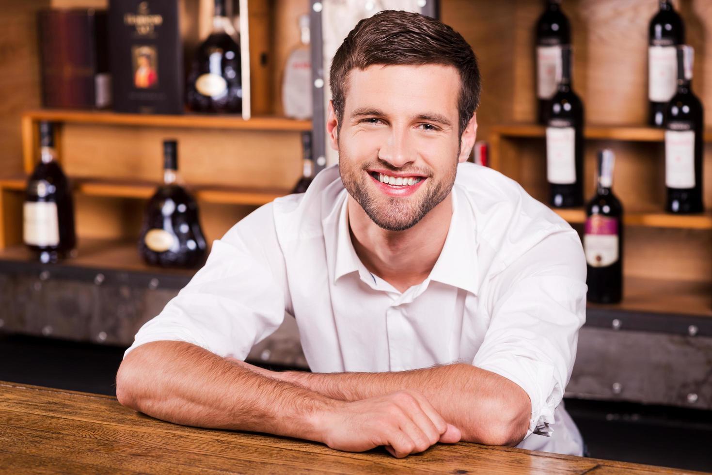 allegro barista. bello giovane maschio barista nel bianca camicia pendente a il bar contatore e sorridente foto