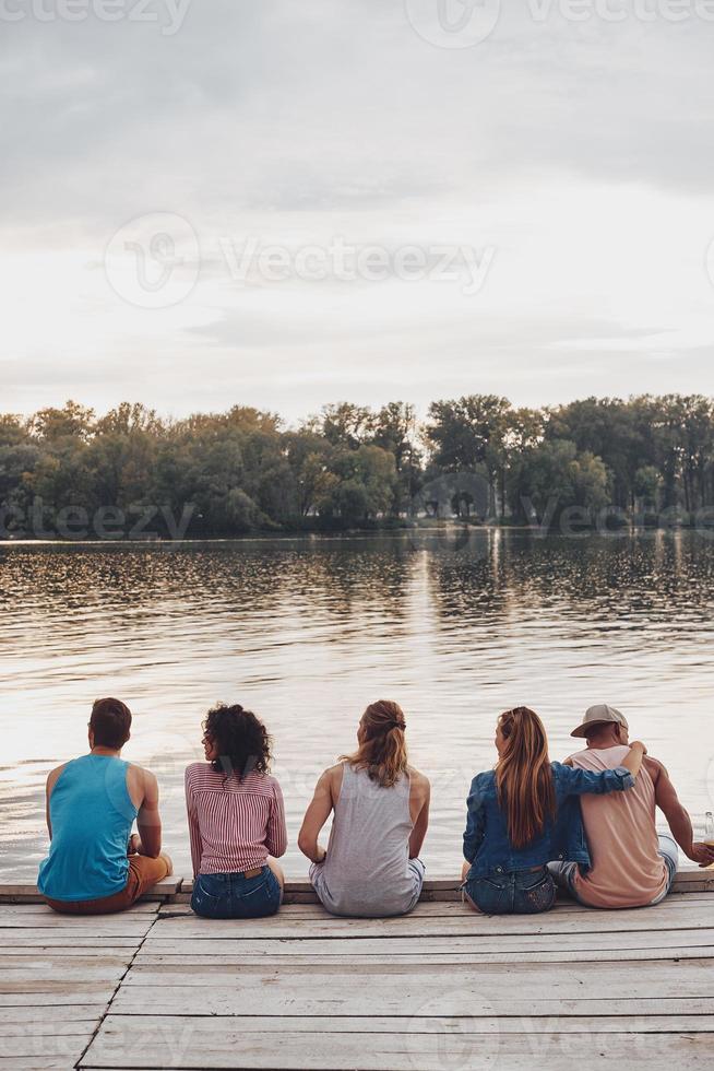 simpatico tempo con gli amici. posteriore Visualizza di giovane persone nel casuale indossare parlando mentre seduta su il molo foto