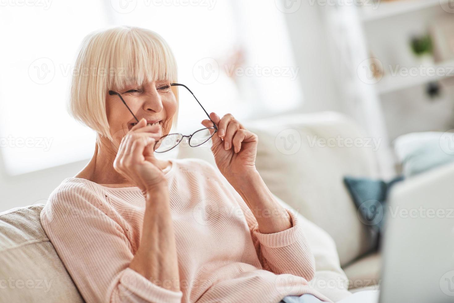 contento nonna. bellissimo anziano donna mettendo su sua occhiali mentre rilassante su il divano a casa foto